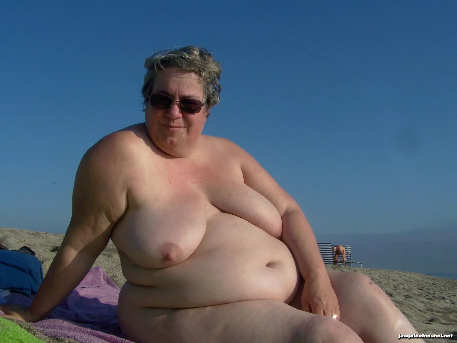 Older nudist with a big belly sitting on the beach