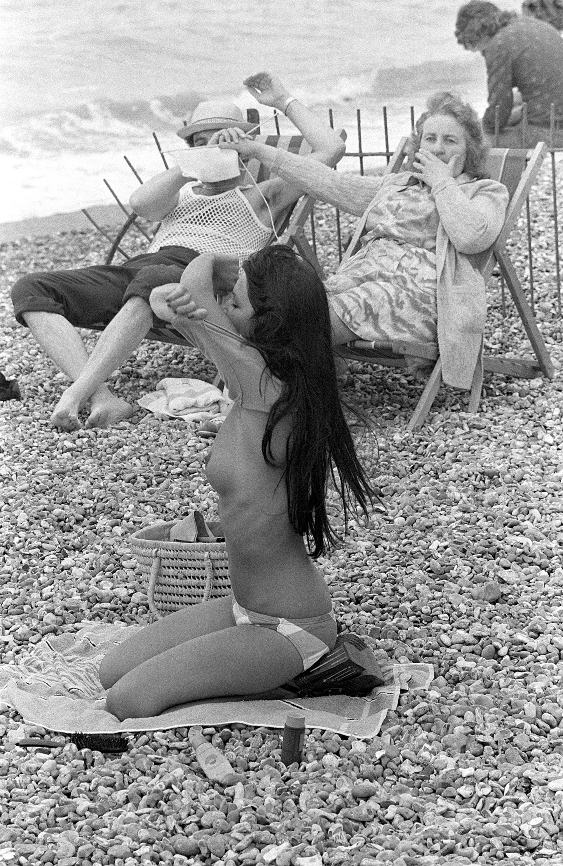 Woman covering man's eyes with her knitting at sight of young woman taking off her top on the beach in France, 1974